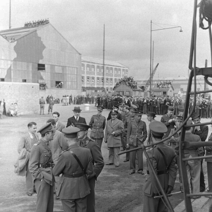 British and American military officers gather along with newspaper correspondents and dignitaries while crowds gather on the roof of buildings in the Harland and Wolff Ltd. shipyard in Belfast.