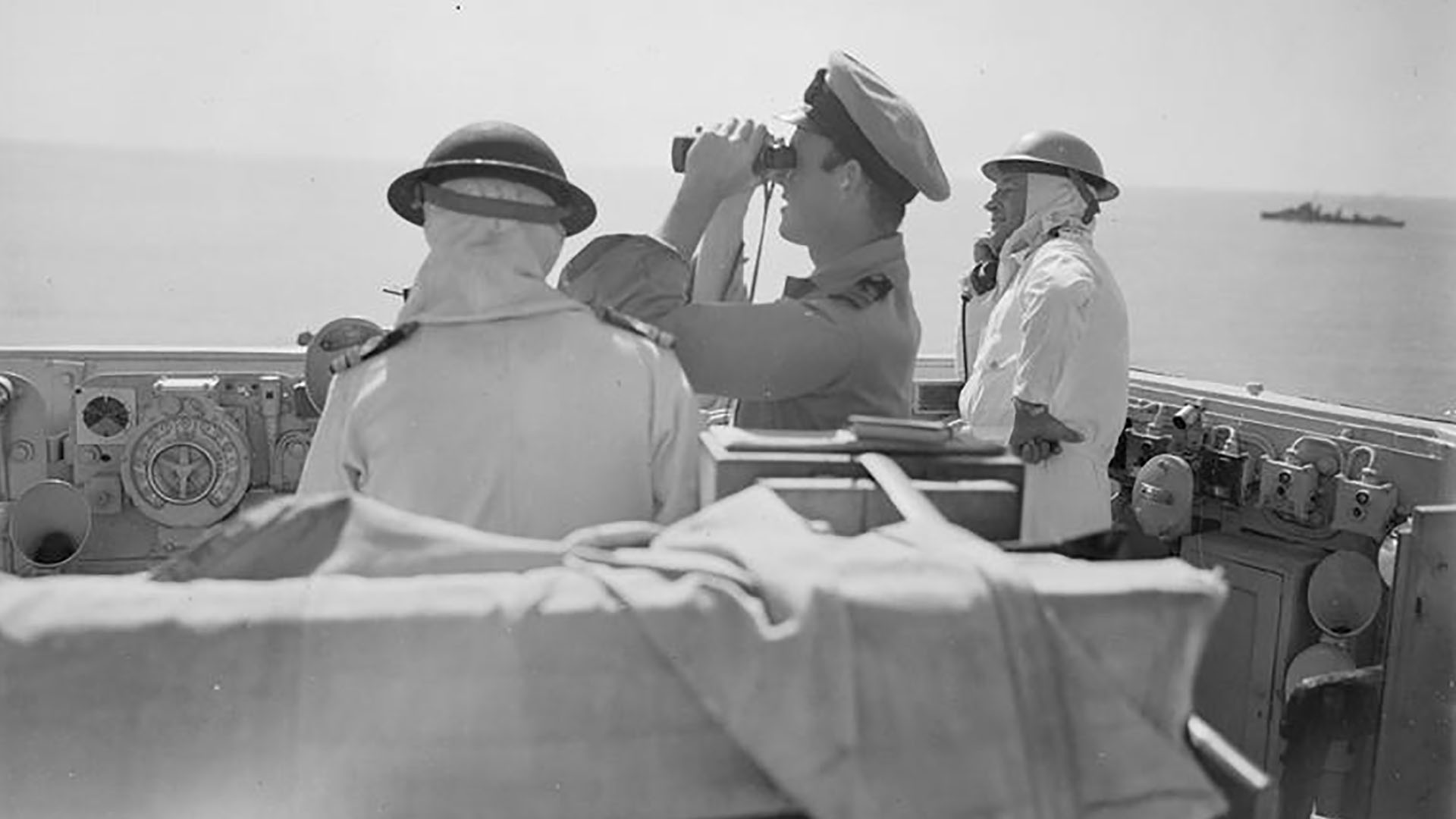 Captain Packer on H.M.S. Warspite directs the bombardment of Reggio during the opening offensive of the Italian campaign. Other crew members wear anti-flash hoods to help prevent sunburn.